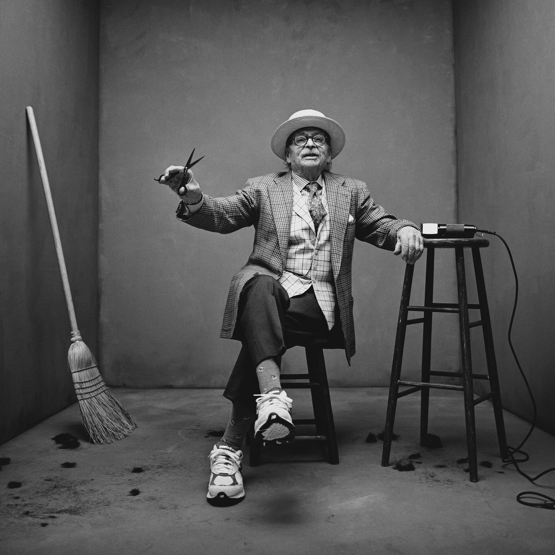 Black and white image of an older craftsman sitting in a stool in a concrete studio backdrop. He is holding a pair of scissors and wearing a pair of MADE in USA sneakers.